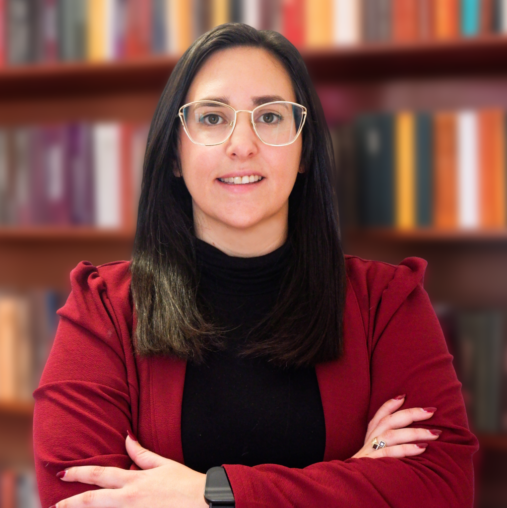 Advogada Carolina Bucete Oliveira, com uma camisola preta e um casaco vermelho, sentada numa cadeira, com uma estante por trás. Na mesa está um livro vermelho.