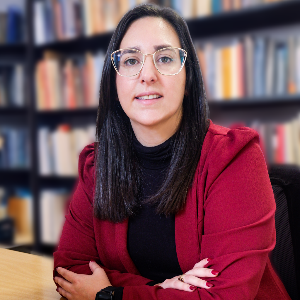 Advogada Carolina Bucete Oliveira, com uma camisola preta e um casaco vermelho, sentada numa cadeira, com uma estante por trás. Na mesa está um livro vermelho.